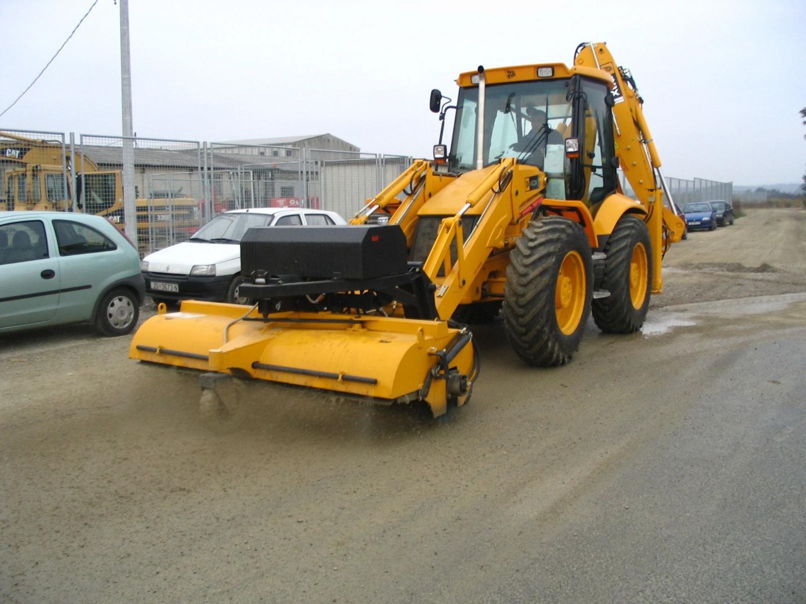 A backhoe loader equipped with a Modularis Drive Loader Sweeper clearing dust and debris from a construction site road.