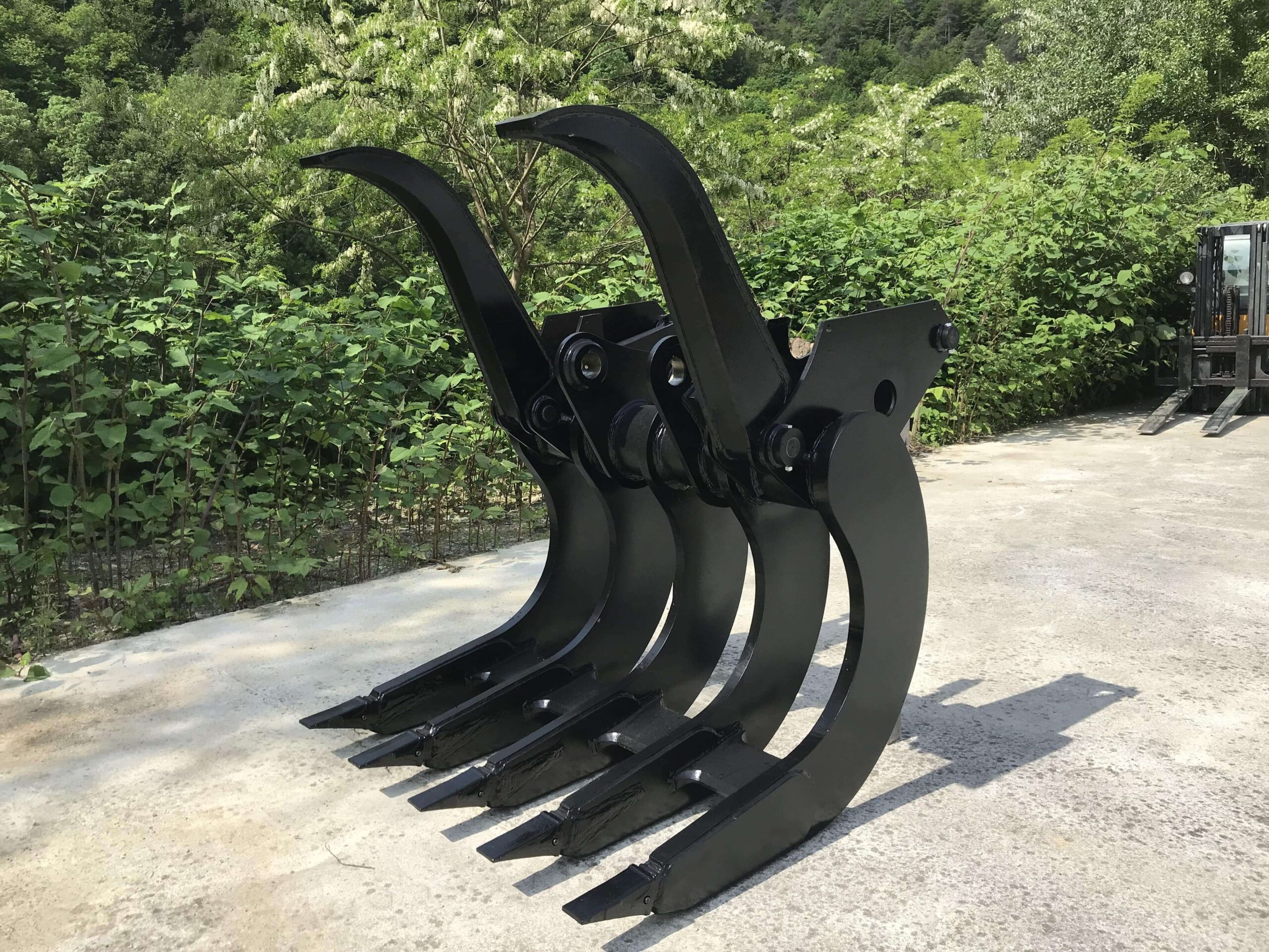 md-rake-25-0-1-min A heavy-duty Modularis Drive Clearing Rake, painted black, standing on a concrete surface outdoors. The attachment features multiple curved tines designed for land clearing, root removal, and debris handling. Green foliage and a partial view of a forklift are visible in the background.