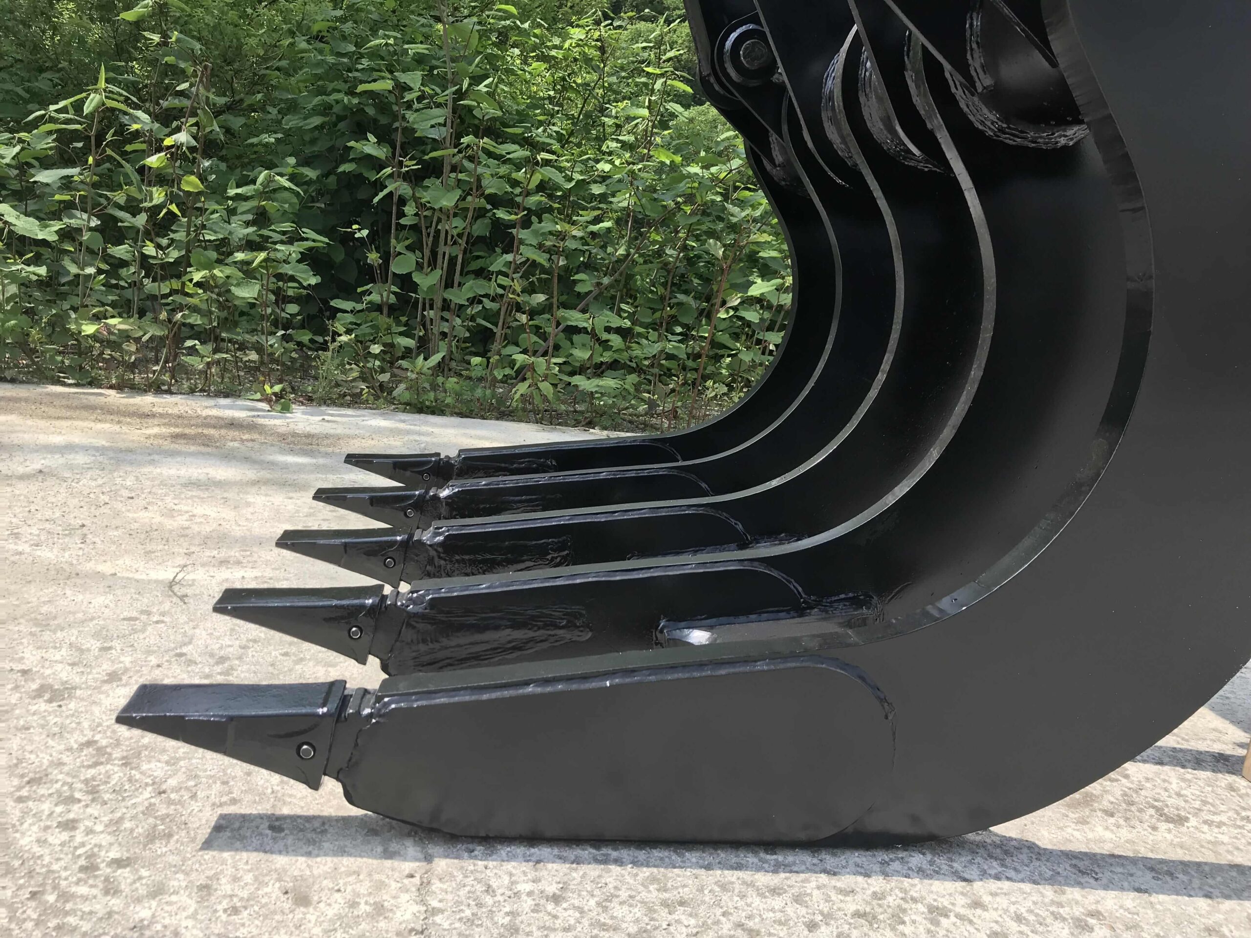 md-rake-25-0-2-min A close-up of the Modularis Drive Clearing Rake, highlighting its sharp, reinforced tines and robust construction. The attachment is designed for efficient ground clearing and material handling. The background consists of dense greenery and a concrete surface.