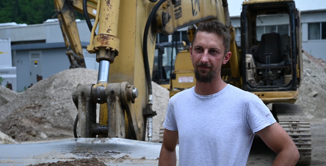 20 Year Testimony Gradbeništvo Gotovnik Construction worker standing next to Caterpillar excavator on construction site with gravel.