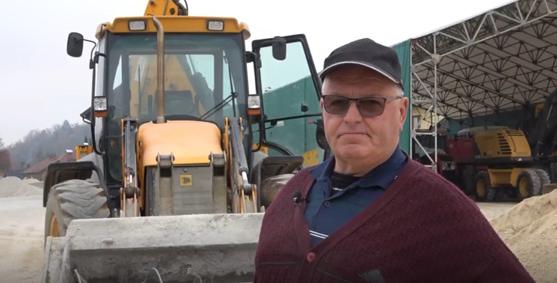 20 Year Testimony Jože Older man standing next to JCB backhoe loader with Modularis Drive concrete mixing bucket attached.